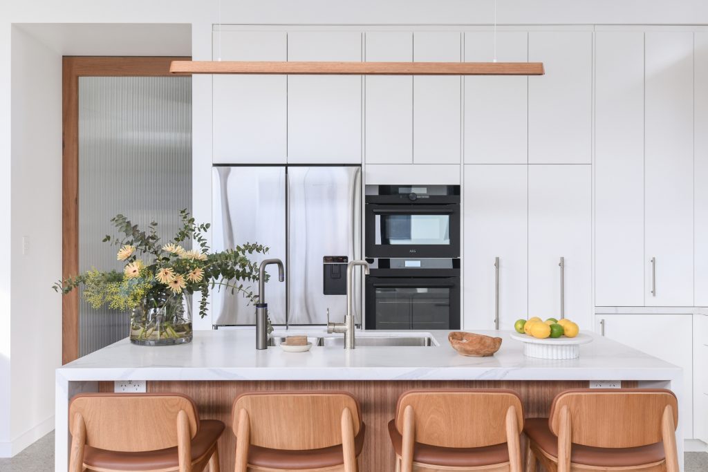 A modern Stanmore House kitchen with white cabinetry, a marble-topped island with four wooden stools, stainless steel appliances, and a vase of yellow flowers.