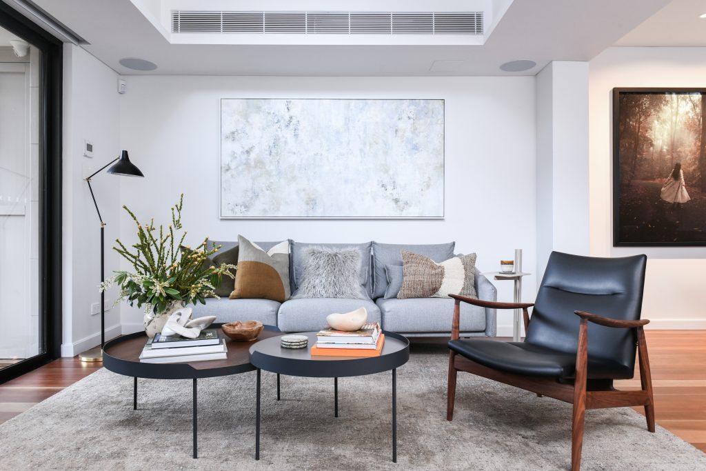 A Victorian terraced living room with a grey sofa, two black coffee tables, a black leather lounge chair, and large abstract wall art.