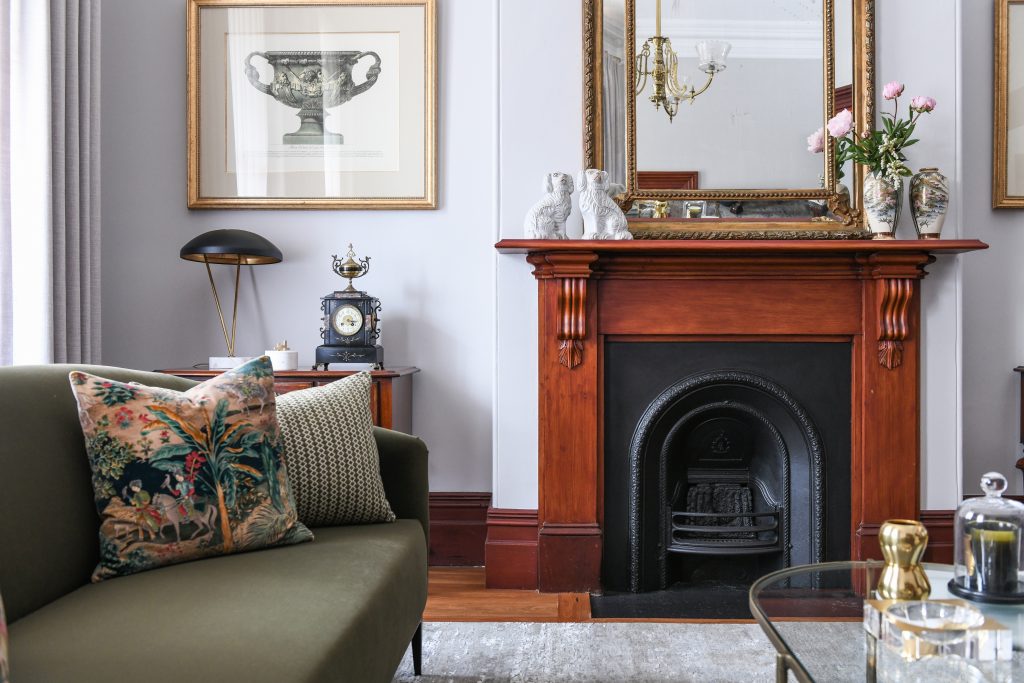 An Inner West Terrace living room with an olive green sofa, a wooden fireplace mantel decorated with two dog figurines and a mirror, and a framed botanical print on the wall.