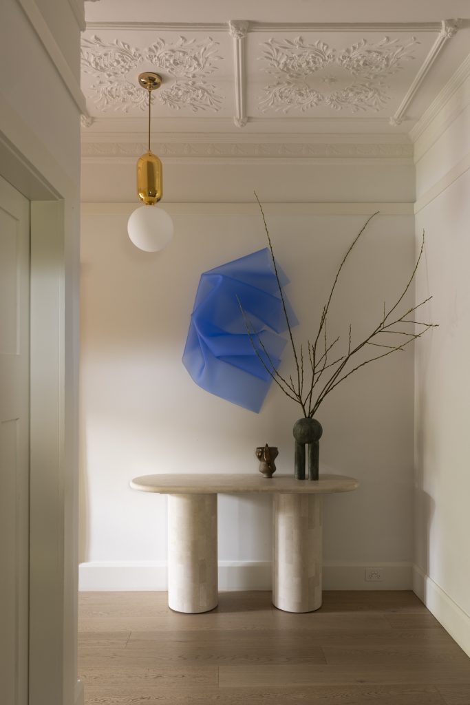 A console table with branches and a sculpture sits beneath blue art and a gold pendant in a Haberfield House interior, framed by ornate ceiling moulding.