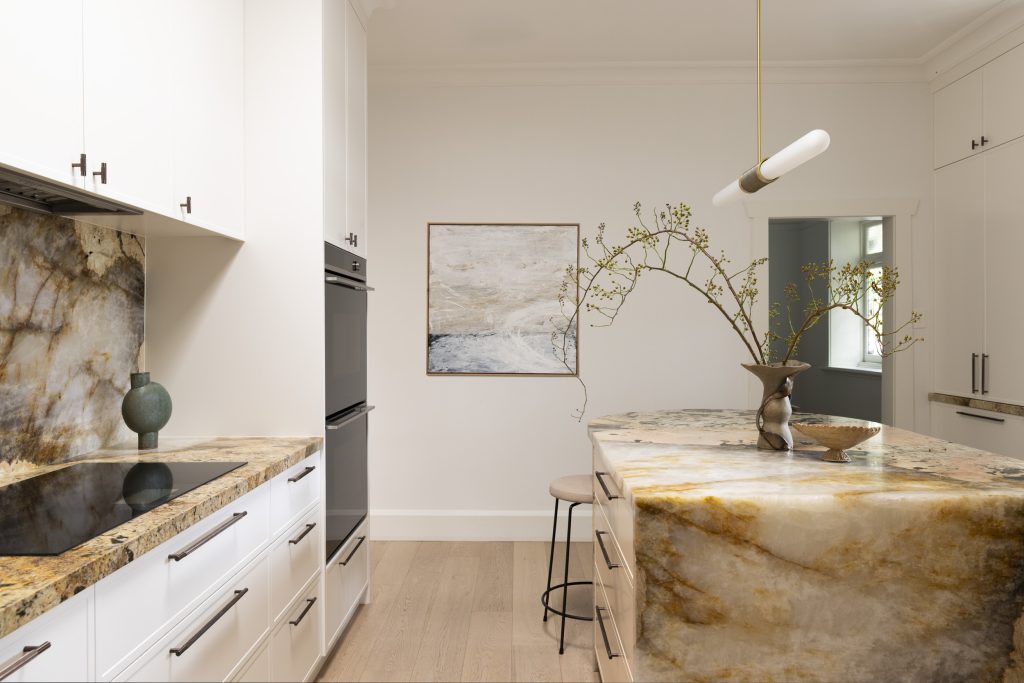 Modern kitchen in a Californian bungalow renovation, featuring white cupboards, a large stone-topped island, a built-in oven, and abstract wall art.