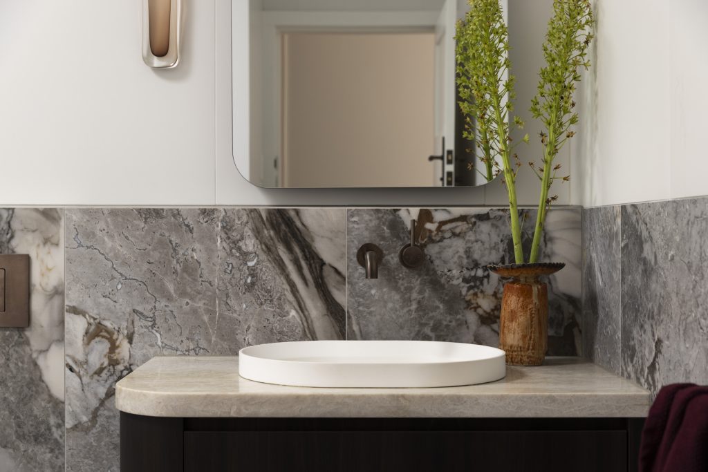A white vessel basin sits on a stone worktop against a grey marble splashback, embodying the style of interior design in Sydney Inner West, with a small vase of greenery to the right.