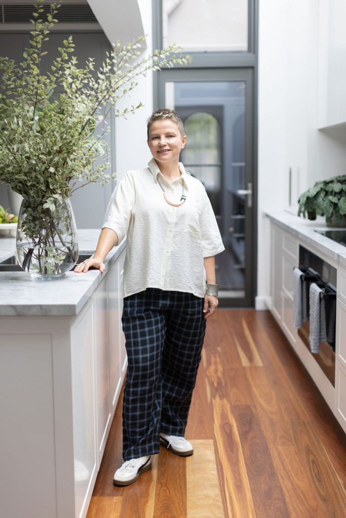 Viktoryia Tsymbal, founder of DOMI Interiors, standing in a white kitchen beside a marble island and a vase of greenery
